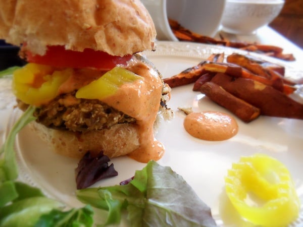 An eggplant veggie burger topped with sliced pickles, tomato, and creamy sauce sits on a plate with sweet potato fries and leafy greens. A dollop of sauce is on the plate beside the burger.