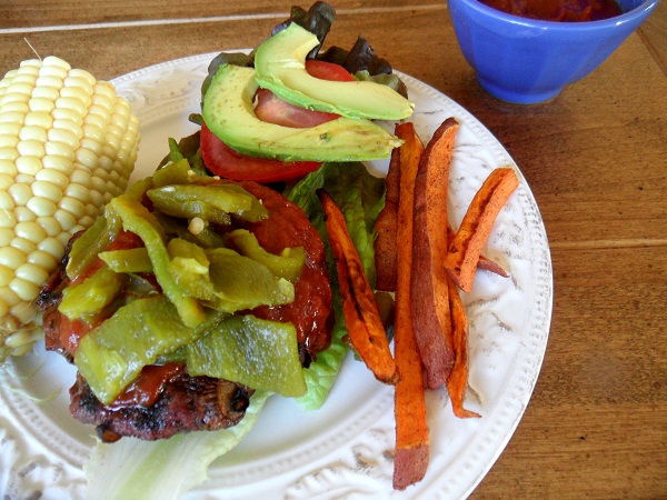 A plate with a grilled burger topped with green chilies, lettuce, tomato, and avocado, served alongside sweet potato fries and half a corn on the cob. A blue bowl with sauce sits in the background.