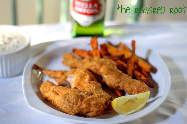A plate of crispy fried fish fillets with sweet potato fries and a lemon wedge, served alongside a small dish of dipping sauce. A blurred bottle and the roasted root text appear in the background, evoking classic baked salmon fish and chips vibes.