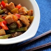 A white bowl filled with turkey stir fry—tender pieces, green beans, red and green bell peppers over rice—rests on a blue textured surface next to a pair of chopsticks.