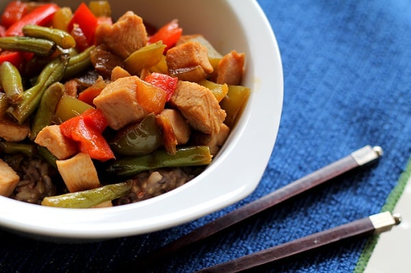 A white bowl filled with turkey stir fry—tender pieces, green beans, red and green bell peppers over rice—rests on a blue textured surface next to a pair of chopsticks.