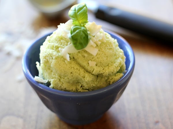 A scoop of basil coconut milk ice cream, garnished with coconut flakes and a fresh basil leaf, served in a small blue bowl on a wooden surface.