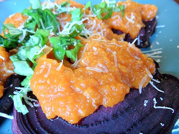 Close-up of Beet Ravioli featuring roasted beet slices filled with orange sweet potato mash, grated cheese, and fresh chopped herbs on a blue plate.