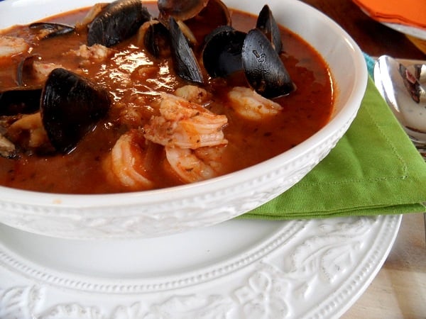 A close-up of a white bowl filled with Seafood Cioppino, featuring shrimp and mussels in a red tomato broth, resting on a green napkin and white plate.