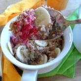 A spoonful of Raspberry Baked Steel Cut Oatmeal topped with sliced bananas and bright red jam is held above a white bowl filled with more oatmeal, banana slices, and jam; a green napkin is underneath the bowl.