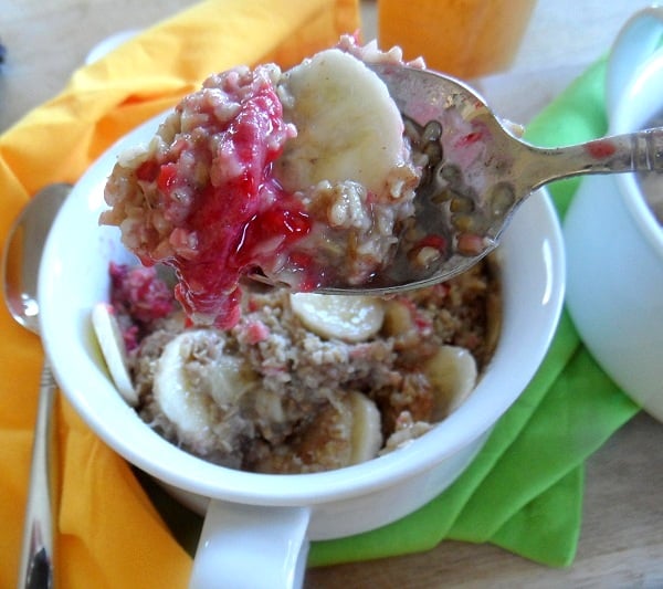 A spoonful of Raspberry Baked Steel Cut Oatmeal topped with sliced bananas and bright red jam is held above a white bowl filled with more oatmeal, banana slices, and jam; a green napkin is underneath the bowl.