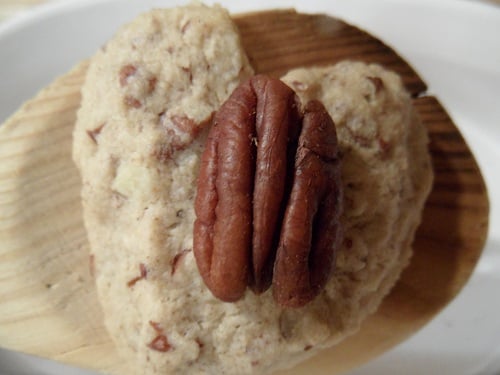 A heart-shaped oat flour shortbread cookie with nut pieces, topped with a whole pecan, sits on a light wooden surface.