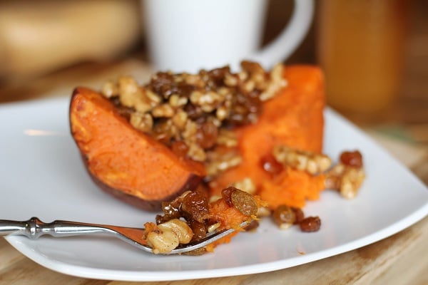 A baked sweet potato half on a white plate, reminiscent of roasted yams, topped with walnuts and raisins. A fork holds a bite-sized portion in the foreground, while a blurred white mug sits in the background.
