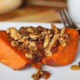 A baked sweet potato half reminiscent of roasted yams is topped with walnuts and raisins, sitting on a white plate with a fork and cup in the background.