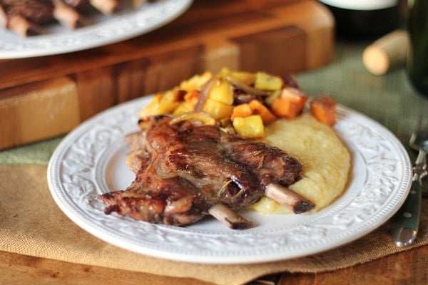 A white plate with Red Wine braised spare ribs, creamy polenta, and a colorful medley of roasted vegetables, set on a rustic table with wooden boards in the background.