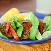 Three fajita lettuce wraps filled with seasoned ground meat, red bell peppers, and onions are served on a blue plate with lime wedges and corn on the cob. A refreshing glass of lemonade sits in the background.