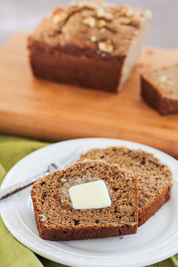 A slice of Vegan Sourdough Banana Bread with a pat of butter on top sits on a white plate, with another slice and the loaf resting on a wooden cutting board in the background.