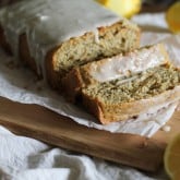 A loaf of gluten free lemon poppy seed bread with white glaze sits on parchment paper on a wooden board. Two slices are cut, and whole and halved lemons are visible nearby.