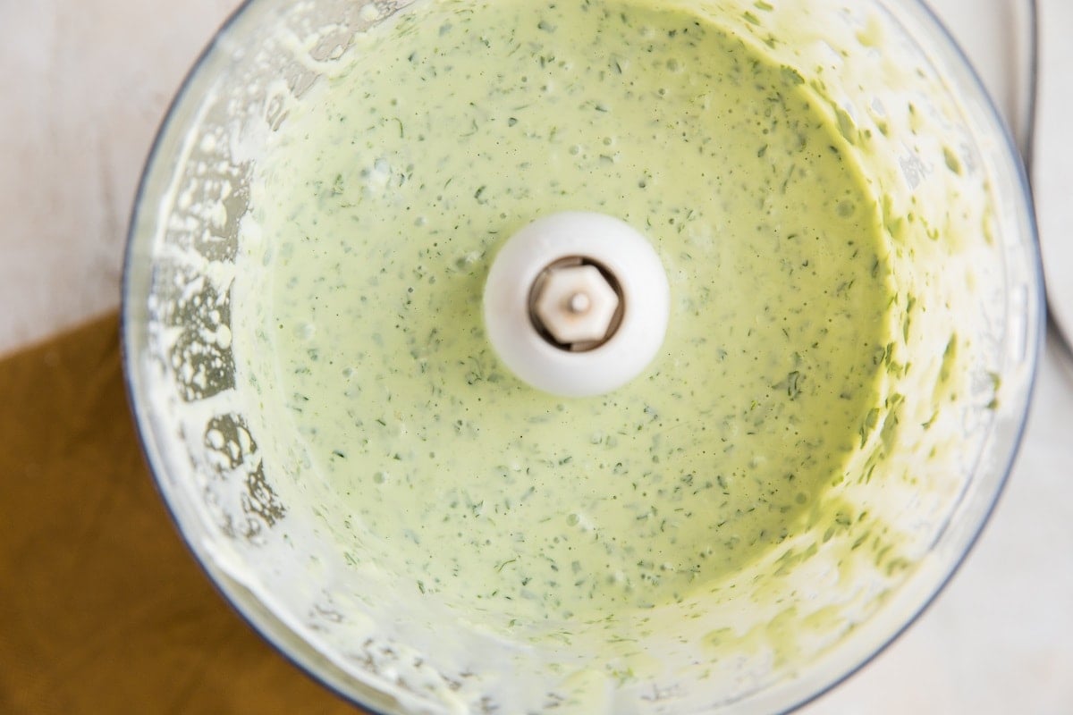 A food processor bowl filled with creamy mayo-free green goddess dressing, likely made with fresh herbs, seen from above with vibrant sauce splatters on the inner sides.
