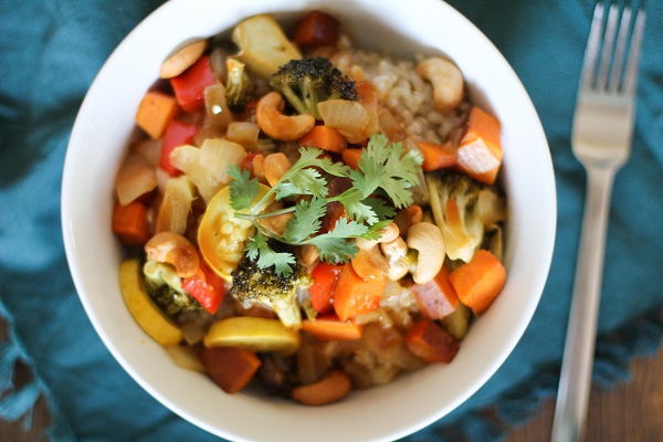 A white bowl filled with colorful roasted vegetables, brown rice, cashews, and topped with fresh cilantro, placed on a green cloth next to a fork.