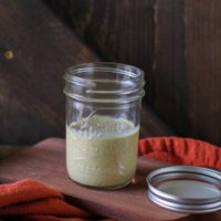 A small glass jar filled with light green kombucha vinaigrette sits on a wooden board next to a jar lid, with an orange cloth draped nearby and a rustic wooden background.