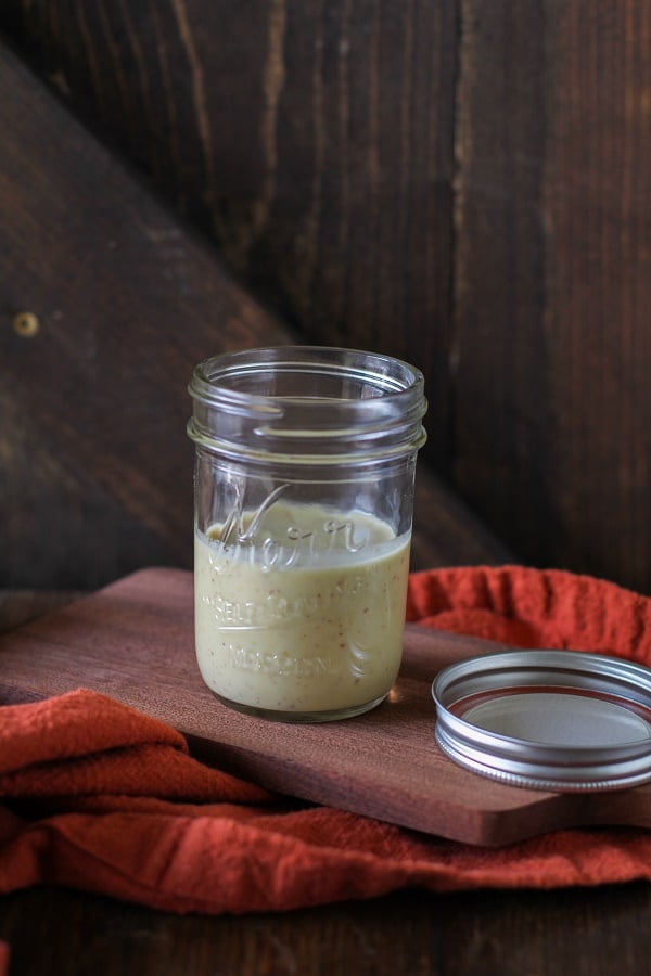 A small glass jar filled with light green kombucha vinaigrette sits on a wooden board next to a jar lid, with an orange cloth draped nearby and a rustic wooden background.
