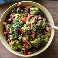 A bowl of salad featuring brown rice bowls with grains, leafy greens, diced vegetables, and generous dollops of green pesto sits on a wooden surface, accompanied by a fork and a blue napkin.