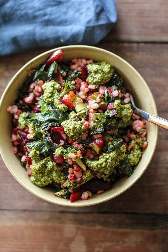 A bowl of salad featuring brown rice bowls with grains, leafy greens, diced vegetables, and generous dollops of green pesto sits on a wooden surface, accompanied by a fork and a blue napkin.