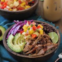A colorful bowl with crock pot Hawaiian pulled pork, sliced avocado, black beans, purple cabbage, lime, and pineapple salsa over quinoa. An iced coffee and another bowl of salsa are in the background.