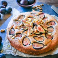 A round almond flour fig cake topped with fresh fig slices and chopped nuts, placed on parchment paper, with whole figs and a bowl of figs in the background.
