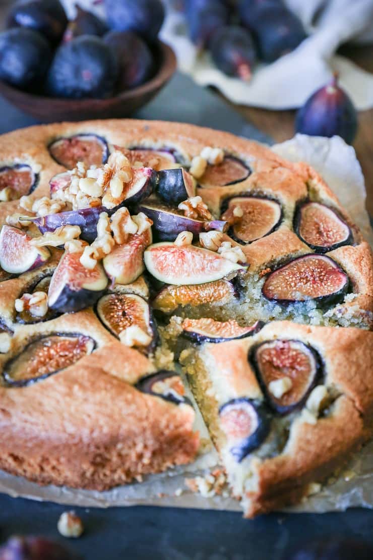 A round almond flour fig cake topped with fresh fig slices and chopped walnuts, with one slice cut out. Whole figs and a bowl of figs are visible in the background.