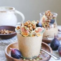A glass jar filled with creamy pumpkin spice chia pudding, topped with granola, sliced figs, and pumpkin seeds, sits on a wooden plate with a spoon, fresh figs, and more seeds scattered around, with another jar in the background.
