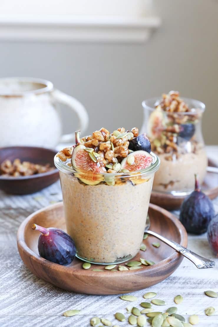 A glass jar filled with creamy pumpkin spice chia pudding, topped with granola, sliced figs, and pumpkin seeds, sits on a wooden plate with a spoon, fresh figs, and more seeds scattered around, with another jar in the background.
