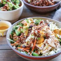 A bowl of crunchy quinoa broccoli cabbage spinach salad with chopped chicken, carrots, and greens, topped with creamy dressing. Other salad bowls and dressing are blurred in the background on a wooden table.