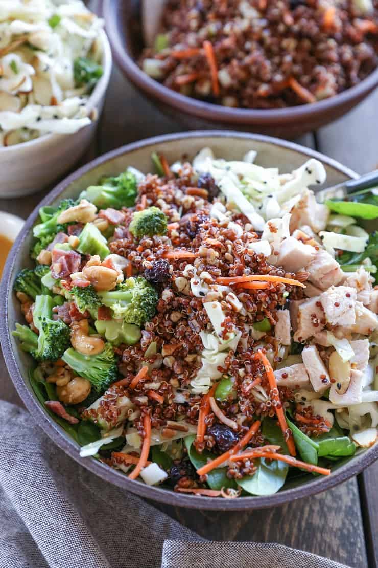 A bowl filled with a colorful, crunchy quinoa broccoli spinach salad featuring chopped chicken, shredded carrots, nuts, and raisins, with other bowls of fresh salad ingredients in the background.