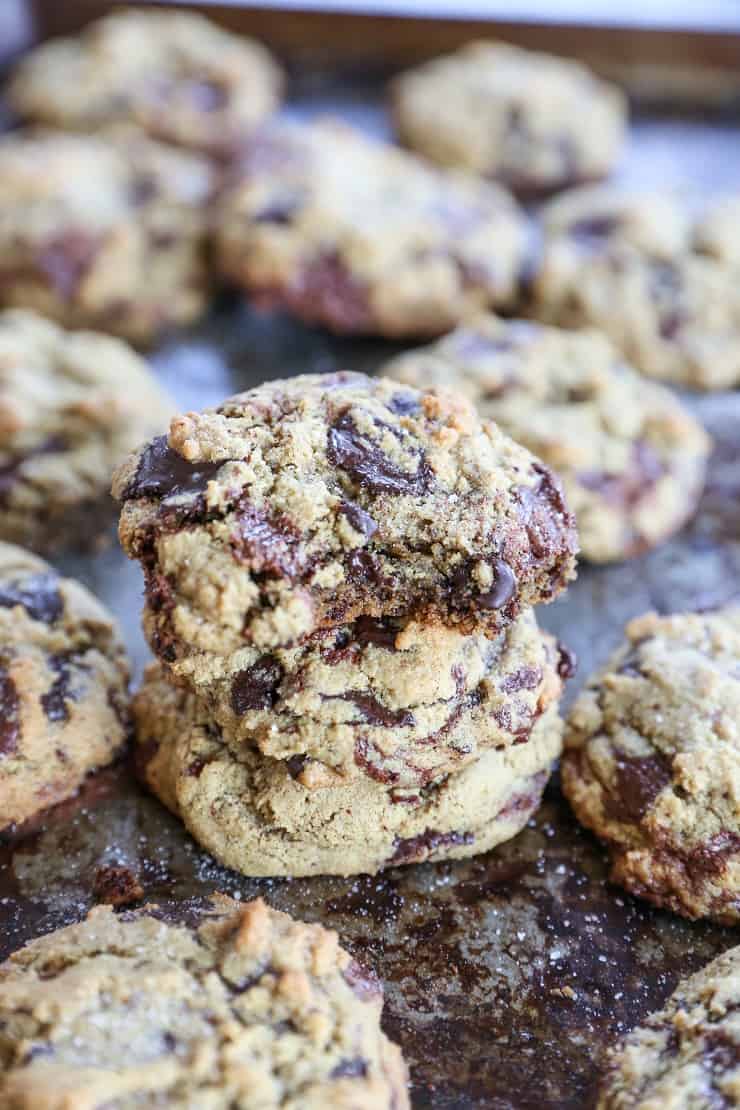 A close-up of two thick, paleo chocolate chip cookies stacked on a baking sheet, surrounded by more cookies in the background. The cookies are golden brown with visible chocolate chunks.
