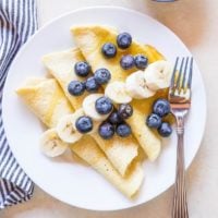 A white plate with folded paleo crepes topped with banana slices and blueberries sits next to a fork. A striped napkin is on the side, and a bowl of fresh blueberries is in the upper right corner.