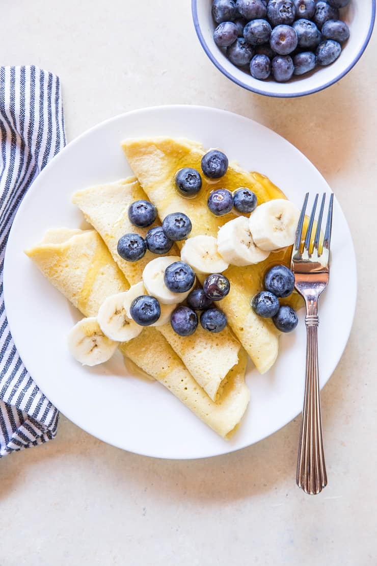 A white plate with folded paleo crepes topped with banana slices and blueberries sits next to a fork. A striped napkin is on the side, and a bowl of fresh blueberries is in the upper right corner.