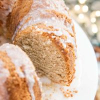 A close-up of a sliced gluten-free eggnog cake with a light glaze on top, displayed on a white plate. The background is softly blurred with twinkling lights, giving a festive feel.