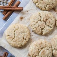 Five golden brown keto snickerdoodles rest on parchment paper, sprinkled with cinnamon sugar. Cinnamon sticks and a gold snowflake decoration are arranged nearby on a light grey surface.
