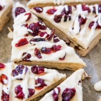 Triangles of iced paleo cranberry bliss bars topped with dried cranberries and drizzled with white icing, arranged closely together on a parchment-lined surface.