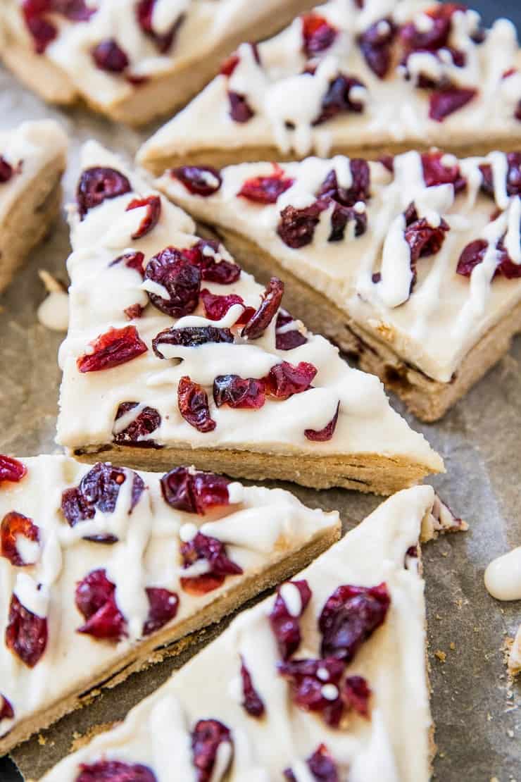 Triangles of iced paleo cranberry bliss bars topped with dried cranberries and drizzled with white icing, arranged closely together on a parchment-lined surface.