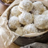 A metal bowl lined with parchment paper holds a pile of paleo Russian tea cakes dusted with powdered sugar, with a small bowl of chopped walnuts and stacked plates in the background.