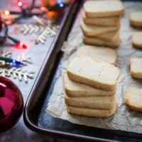 A stack of 3-Ingredient Paleo Shortbread cookies on a baking tray lined with parchment paper, surrounded by festive holiday decorations and colorful string lights.