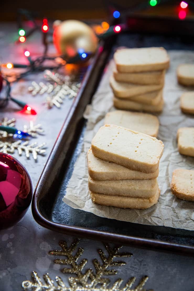 A stack of 3-Ingredient Paleo Shortbread cookies on a baking tray lined with parchment paper, surrounded by festive holiday decorations and colorful string lights.