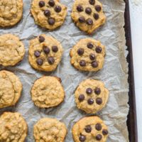 A baking tray lined with parchment paper holds twelve homemade paleo banana bread cookies, some topped with chocolate chips and others plain, arranged in neat rows.