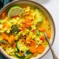 A bowl of vibrant turmeric vegetable soup with rice, carrots, zucchini, broccoli, and red pepper flakes, garnished with a lime wedge. A spoon rests in the bowl and a small dish of turmeric is nearby.