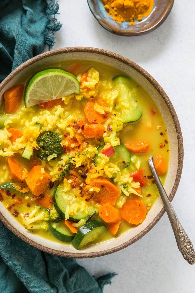 A bowl of vibrant turmeric vegetable soup with rice, carrots, zucchini, broccoli, and red pepper flakes, garnished with a lime wedge. A spoon rests in the bowl and a small dish of turmeric is nearby.