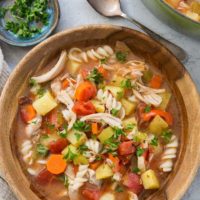 A wooden bowl filled with Sicilian chicken soup, brimming with pasta, potatoes, carrots, tomatoes, and fresh herbs. A spoon, a napkin, and a small dish of chopped parsley sit nearby, with a pot of soup in the background.