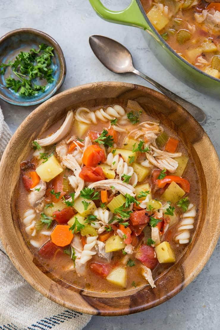 A wooden bowl filled with Sicilian chicken soup, brimming with pasta, potatoes, carrots, tomatoes, and fresh herbs. A spoon, a napkin, and a small dish of chopped parsley sit nearby, with a pot of soup in the background.