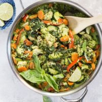 Skillet of green curry with vegetables, chicken, and rice, with a napkin and a bowl of fresh lime wedges to the side. Ready to serve.