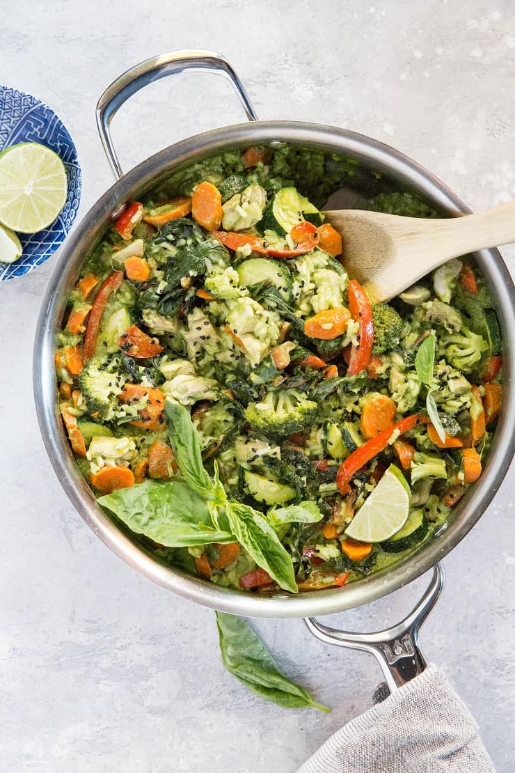Skillet of green curry with vegetables, chicken, and rice, with a napkin and a bowl of fresh lime wedges to the side. Ready to serve.