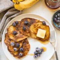 A plate of 3-Ingredient Banana Pancakes is topped with blueberries, a pat of butter, and syrup, next to a fork. Fresh blueberries, two bananas, a jar of syrup, and a napkin are in the background.