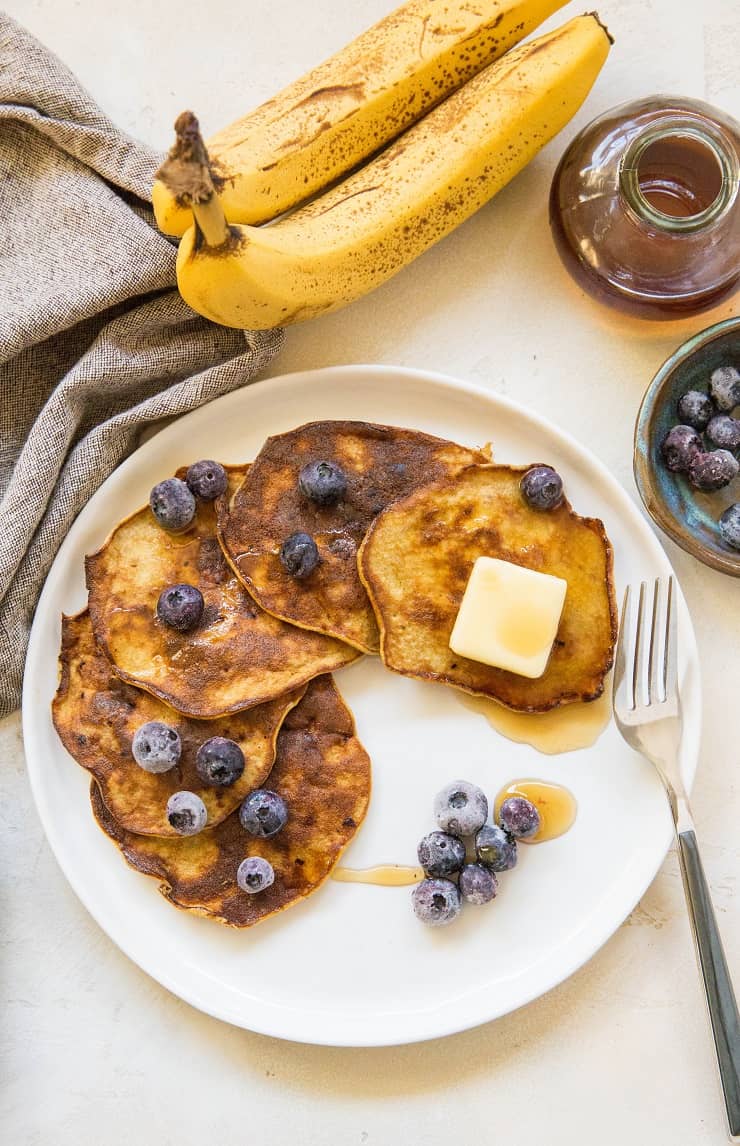 A plate of 3-Ingredient Banana Pancakes is topped with blueberries, a pat of butter, and syrup, next to a fork. Fresh blueberries, two bananas, a jar of syrup, and a napkin are in the background.