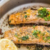 Two pan-fried salmon fillets in a metal pan, garnished with chopped herbs and spices, with a halved lemon placed beside the fillets.